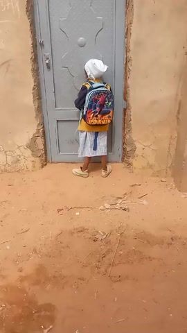 A young child with a white head covering and a Spiderman backpack stands facing a gray metal door set in a textured, earthen wall. The child is wearing a yellow shirt, a checkered skirt, and tan shoes. They are reaching for the door's knob. The ground is sandy and appears dry. The surrounding walls show signs of age and weathering.