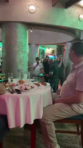 In a brightly lit restaurant, a group of six men and a boy gather around a small drum set. The boy, in a white polo shirt, records the scene with his phone. One of the men, bald and wearing glasses and a patterned shirt, plays the drums. The other men surround him, clapping and gesturing, seemingly enjoying the performance. A table laden with food sits in the foreground, partially obscuring the view. The restaurant's interior features columns and a fish tank, adding to the atmosphere.
