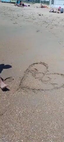A person draws a heart in the sand on a beach. Other people are visible relaxing in the background. The waves are approaching the shore.