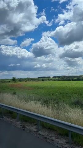 The view from a vehicle moving along a road reveals a pastoral scene. A green field dotted with patches of brown stretches towards the horizon, framed by a cloudy blue sky. In the foreground, a grey metal guardrail lines the roadside. The clouds overhead are varied in size and texture, ranging from fluffy white to darker, more ominous shades.