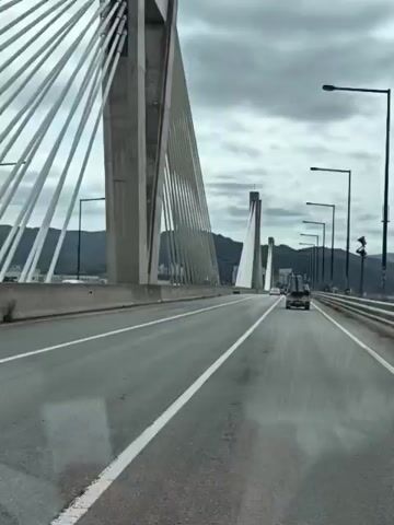 This video shows a view from a vehicle driving across a bridge in Sejong, South Korea. The bridge is a modern cable-stayed bridge with tall concrete towers and numerous cables. The road surface appears wet, suggesting it might be raining or has recently rained. The sky is overcast with dramatic clouds.

In the foreground, the side of a white truck is visible, indicating the perspective of the camera. Further ahead on the bridge, other vehicles, including trucks and cars, can be seen driving in the same direction. The sides of the bridge have guardrails, and lampposts are spaced along the roadway.

Beyond the bridge, the landscape is hilly and covered in lush green vegetation. In the distance, mountains are visible. The overall mood of the video is somewhat somber due to the cloudy weather, but the impressive architecture of the bridge and the natural beauty of the surrounding landscape are prominent. No text is visible in the video. The time of day appears to be daytime, given the ambient light.