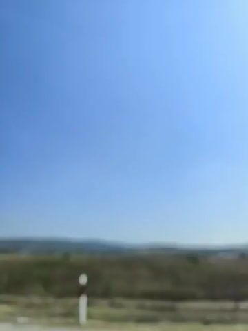 The video captures a view from a vehicle moving along a road. The landscape is rural, with fields and rolling hills under a clear blue sky. A metal guardrail runs along the side of the road in the foreground. At one point, the underside of a concrete overpass is visible, casting a shadow. The video appears to be shot during daylight hours, suggesting it is daytime. There are no people or significant human activity explicitly shown in the frame. The overall impression is one of travel through an open, natural environment.