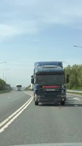 The video shows a view from the inside of a car traveling on a highway. A blue Scania 94G 300 truck is in the foreground in the same lane, driving ahead of the car. Other vehicles, including another truck, can be seen further ahead in the distance. The highway is marked with white lines and has streetlights along the side. There is green vegetation on the side of the road and a partly cloudy sky above. The camera is stable and pointed toward the front. The atmosphere seems to be daytime and the weather is sunny.