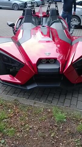 A red Polaris Slingshot is parked on a brick-like surface with grass and leaves in the foreground. A man in a dark shirt and blue jeans stands to the right of the vehicle, facing away from the camera, with his right hand on his hip. Other cars are visible in the background.