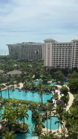 This video shows a high-angle, static view of a resort with multiple buildings and swimming pools surrounded by lush tropical vegetation. In the foreground, several expansive swimming pools are visible, filled with turquoise water and surrounded by lounge chairs and palm trees. Some people are seen in the pools and on the pool decks, relaxing and swimming. In the background, two large, multi-story hotel buildings dominate the skyline. The building on the right is a cream-colored high-rise with many balconies. The building on the left is a more uniquely designed structure with a curved, stepped facade and numerous openings. The sky above is partly cloudy. The overall impression is one of a luxurious tropical vacation destination.
