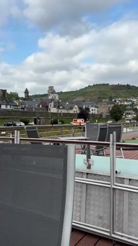 The video shows a view from a deck with chairs looking out at a town nestled in a valley. The town has a church with a tall tower and other buildings. A road runs in front of the town, and a white tour bus is driving on it. There are also cars and an ambulance visible on the road. The town is surrounded by green hills, some of which are covered in vineyards. The sky is cloudy with patches of blue.