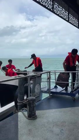 The video shows three people unloading luggage from a boat onto a pier. The people are wearing red shirts and life vests. One person unloads a rolling suitcase from a blue cart. The other two people place it, along with a backpack, onto a pile of luggage. The luggage is a mix of black, blue, and purple rolling suitcases. The pier has a roofed area with decorative railings and is adjacent to the water. The water is choppy, and the sky is overcast. A motorcycle is visible in the background. The sound of the ocean wind is prominent.