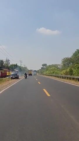 A vertical video shows a highway in daylight. The road has two lanes with yellow dividing lines. On the left side of the road, there is a black car parked, and a person on a motorcycle is visible. Further down the road, there are a few other vehicles, including trucks and motorcycles. The right side of the road is bordered by a metal guardrail and lush green trees. The sky is blue with a few white clouds, and the overall atmosphere is sunny.