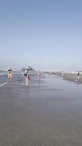 The video shows a beach scene in Blankenberge, Belgium. Several people, including children and adults, are enjoying the beach. Some are wading in the shallow water, while others are walking along the shore. A pier extends into the ocean in the background. The weather appears to be sunny with a clear blue sky, suggesting it is daytime. The overall atmosphere seems relaxed and pleasant, with people engaging in typical beach activities. There is no visible text in the video.