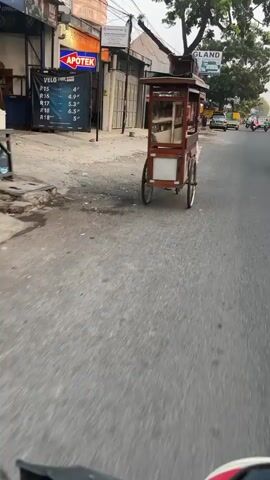 The media file captures a street scene in Larangan, Indonesia. The primary subject is a vendor pushing a wooden cart with a glass display case. The cart appears to be selling food or drinks, with a shelf visible inside. The vendor is moving down a paved road, away from the camera.

The scene is an urban environment with buildings lining the street. One building has a prominent sign for "APOTEK" (Pharmacy) in English and Indonesian. Another building has a sign with "GLAND" visible, possibly an advertisement or part of the building's name. There's a sign on a pole with a list of wheel sizes (R15, R16, R17, R18) and corresponding measurements in inches (4", 4.9", 5.3", 6.3", 5").

The time of day appears to be late afternoon or early evening, as the lighting suggests the sun is low, casting some shadows. The weather seems overcast or hazy. The road is relatively empty, with a few other vehicles, including motorcycles, visible in the distance. The overall mood is that of everyday life in a busy street, with the vendor engaged in their work. The ground shows signs of being a well-trafficked area, with some debris present. The camera angle is from a low perspective, possibly from another vehicle or a person walking.
