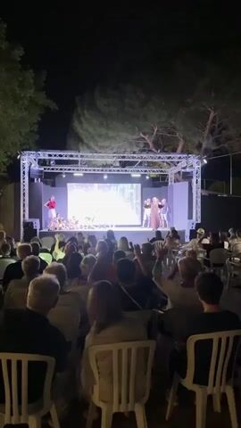This is a vertical video taken at night from behind a seated audience looking towards a stage. The audience members are facing away from the camera, and many have their arms raised. On the stage, there is a large screen displaying an image or video, and in front of it, several performers are standing. A metal truss structure supports lighting and the screen. The environment appears to be outdoors, with trees visible behind the stage. The overall atmosphere suggests an evening performance or event.
