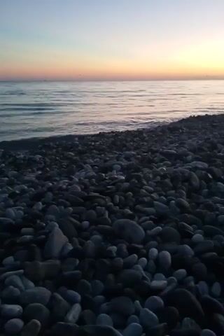 This is a vertical, low-angle, close-up video of a rocky beach. The camera is angled downwards towards the rocks, with the ocean and sky to the left. The rocks are dark gray and vary in size, tightly packed together. The ocean is calm and reflects the orange and yellow hues of the sunset. The sky is a gradient of purple and orange. The lighting suggests it is dusk. There are no people or animals visible in the frame. The overall impression is one of natural serenity.