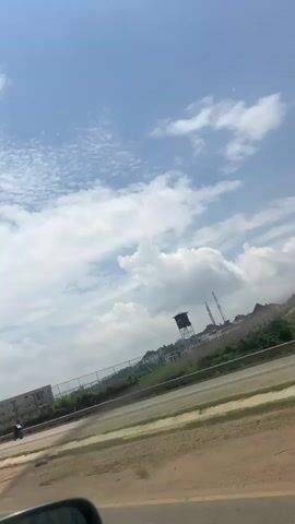 The video captures a view from a moving vehicle on a bright, cloudy day. In the foreground, the edge of a car mirror is visible on the lower left. The road is in the mid-ground, with a clear lane marker and a patch of dry grass. Beyond the road, a fence runs along a gentle incline. On the other side of the fence, there are buildings, some appearing to be residential, and a tall structure resembling a water tower with a crane nearby. A person walks along the road on the left side, and a motorcycle is also visible. The sky is predominantly blue with scattered white clouds, some of which are large and puffy, while others are smaller and wispy. The camera angle is tilted, giving a dynamic perspective of the passing scenery.