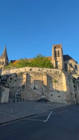 The video presents a panoramic view of a historic town, beginning with a majestic stone church on an elevated platform. The church, featuring a tall spire on the left and a prominent tower with a clock face on the right, is constructed from light-colored stone. Its architecture suggests a Gothic or Romanesque style, with flying buttresses visible along its side. A substantial stone retaining wall forms the base of the elevated church complex, with green foliage growing atop it.

The camera then pans right, revealing a cobblestone street in the foreground transitioning to an asphalt road that curves alongside the church. This road is flanked by high stone walls. The pan continues to show a broader street leading into a town square or courtyard. On the left side of this entrance, a stone gatepost is visible with the word "Mairie" (French for City Hall) carved into it, indicating a municipal building nearby. Surrounding the square are more old stone buildings with distinctive red-tiled roofs and multiple chimneys, bathed in the same warm, golden sunlight.

The sky is clear and blue throughout the video. The long shadows cast by the buildings and walls suggest either early morning or late afternoon light. No people or vehicles are visible, contributing to a serene and timeless atmosphere. The scene is set in what appears to be a well-preserved historic village, likely Saint-Leu-d'Esserent, France. The video concludes by panning back left, retracing the view along the street adjacent to the church.