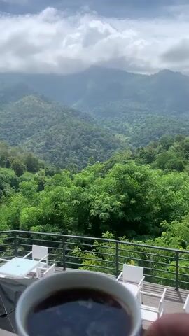 A person's hand holds a white coffee cup filled with dark liquid. The cup is in the foreground, slightly blurred. In the background, a lush green landscape of rolling hills and mountains is visible under a cloudy sky. Some outdoor furniture, including white tables and chairs, is present on a balcony or patio below the mountain view. The video is a close-up shot of the coffee cup with the scenic view beyond.