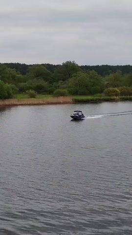 A motorboat speeds across a dark, rippled body of water. The boat is dark blue, with a small cabin and what appears to be two people inside. It leaves a white wake as it moves. The far shore is lined with green trees and some tall reeds. The sky is overcast and gray. The camera seems to be positioned at an elevated viewpoint, possibly on a bridge or hill, overlooking the scene.