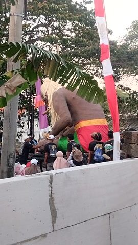 This is a vertical video recording that shows a procession or event involving a large effigy, possibly a giant puppet or statue, decorated with red and white banners. The effigy appears to be human-like with blonde hair and is draped in a yellow and red cloth. A crowd of people, many wearing black t-shirts and some wearing headscarves, are gathered around the effigy, some of whom appear to be walking or dancing. The environment suggests an outdoor setting with trees and a partially visible concrete structure in the foreground. There are also some colorful banners hanging in the background. The lighting indicates it is daytime, possibly late afternoon.
