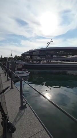 A vertical video captures a wide shot of a modern building on the waterfront with a large, sleek, multi-story structure that appears to be a yacht or a ferry. The building features extensive glass panels, reflecting the sky and clouds. In the foreground, a metal railing lines the edge of a stone walkway. To the left of the frame, a smaller white boat with red trim is docked. The water is calm and reflects the sky, the building, and the sun, which is prominently visible high in the sky with some clouds. The scene suggests a bright, possibly daytime, setting at a marina or harbor.