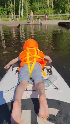 A young person wearing an orange life vest and grey shorts is lying face down on a paddleboard on a lake. Their arms are spread out to the sides, and their feet are pointing away from the camera. A bottle of water is strapped to the paddleboard near their right leg. In the background, several wooden docks are visible, with other people standing and walking on them. The water is dark and reflects the trees and sky. The sun is shining brightly, casting shadows on the paddleboard. The video appears to be filmed from a slightly elevated perspective behind the person on the paddleboard.