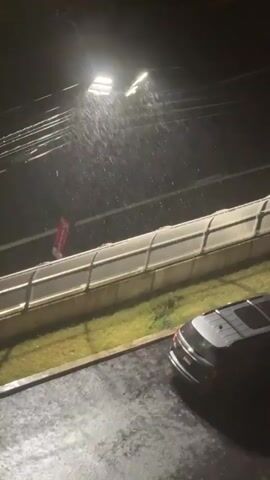 A dark, nighttime scene depicts heavy rain falling under a bright streetlamp. The camera is positioned looking down at an angle. In the foreground, a wet, asphalt surface is visible, with a black SUV parked on the right side. Beyond the asphalt, a grassy area is bordered by a beige and white chain-link fence with a concrete base. Behind the fence, a road or track runs horizontally across the frame, with what appears to be a red sign with white text visible on the left side. Power lines are strung across the upper portion of the image, silhouetted against the dark sky. The rain is clearly visible as streaks falling from the sky.