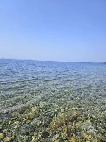 The video shows a clear, shallow body of water with a rocky seabed. The water is calm and reflects the bright blue sky. On the right side of the frame, a rocky cliff covered with green vegetation rises from the water. The top of the cliff appears to have some ancient stone ruins. The perspective is low, suggesting the camera is close to the water's surface. The video pans slowly to the right, revealing more of the coastline and the ruins. There are no people or animals visible in the video.