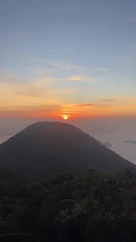 The video shows a time-lapse of a sunrise over a mountain range. The sun is a bright orange ball just above the horizon, casting a warm glow across the sky. The sky is a gradient of blues, oranges, and yellows, with a few wispy clouds. The mountain range is silhouetted against the colorful sky, with lush green trees in the foreground. The water is visible in the distance, reflecting the colors of the sunset. The video pans slightly to the right as the sun rises higher in the sky.