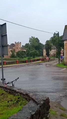 The video shows a slightly overcast day, with a wet road indicating recent rain. In the foreground, on the left, a bicycle is leaning against a low stone wall. The road curves gently, leading towards buildings on the right. One building, with peeling yellow paint and a dark, tiled roof, appears to be old and somewhat neglected. A blue road sign is mounted on its wall, with white text and arrows indicating directions to "AIRUTE 1.5", "UPSIDELE 3.3", and "RUNDALE 12.9". Further down the road, more buildings and trees are visible, and on the left, a castle-like structure with turrets can be seen in the distance. The overall atmosphere is quiet and somewhat melancholic due to the weather and the aged appearance of the buildings.