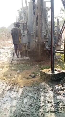In a medium shot during daylight, a muddy ground covers the foreground. A man, wearing a tank top with the number "31" and shorts, operates a large drilling machine. The machine, positioned in the center, is actively drilling, with debris and muddy water spraying around the drill site. The man stands on a platform attached to the machine, facing it while manipulating controls. To the right of the machine is a metal structure or frame, partially obscuring the view. The background includes sparse trees and a building in the distance, suggesting a rural or semi-rural environment.