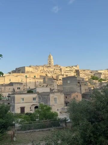 This is a video of a historic Italian city. The city is built into a hillside and features stone buildings with terracotta roofs. A tall bell tower is visible in the distance, rising above the city. The sky is clear and blue. The video appears to be filmed during the day. The camera is panning slightly from left to right, showcasing the architecture of the city. There are no people visible in the video. The overall impression is one of ancient history and timeless beauty.