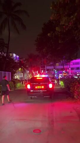 A police SUV with flashing red and blue lights is parked on a street at night. The SUV has the words MIAMI BEACH POLICE written on the back. Several people are visible on the sidewalk in the background, some of whom are walking and interacting. There are palm trees and buildings with neon lights on in the background. The overall scene appears to be a busy urban street during nighttime.