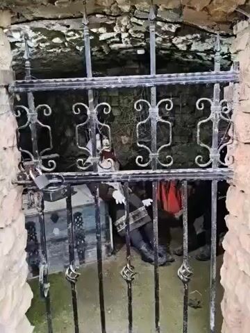 The video shows a dimly lit, rocky interior, possibly a cellar or cave, with a barred gate in the foreground. Inside, behind the gate, a mannequin dressed in traditional clothing, including a headscarf, vest, and patterned skirt, is seated. The mannequin is positioned next to an old phonograph and a chest. Cobwebs are visible, adding to the eerie atmosphere. To the right of the first mannequin, another figure in black clothing and boots is partially visible. The scene appears to be staged, possibly as a display or exhibit.