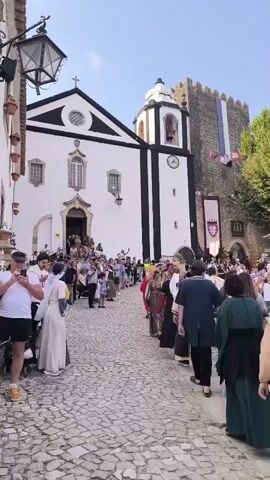 The video opens on a cobblestone street lined with historic buildings, including a white church with black trim and a bell tower. A large crowd of people, many dressed in medieval or Renaissance-style attire, are gathered and appear to be in a procession or waiting for an event. Flags hang from the buildings, suggesting a festival or historical reenactment. The camera pans across the scene, showing individuals in the foreground and mid-ground, some interacting with each other, while others are observing the surroundings. The atmosphere appears to be lively and festive, with a sense of community and historical appreciation. The video then shifts to a different part of the same event, showing more people in costume mingling in a courtyard area with a statue and flowering bushes. The lighting suggests it is daytime.