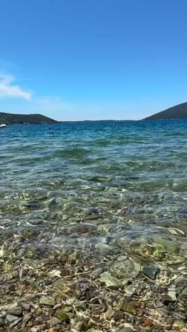 The scene captures a clear, bright day at the seaside. The camera is positioned low, at the edge of a rocky beach. The foreground is dominated by gray and white pebbles, partially submerged by the clear turquoise water. Ripples on the water's surface allow one to see the rocks beneath. The horizon reveals a bright blue sky with only a few faint clouds. In the distance, there are dark, hilly landmasses on either side of the frame. There are a few buoys, and one boat can be seen far away. There are no clear signs of human activity.