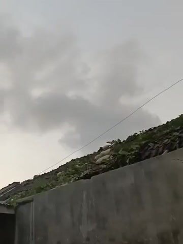 The video shows a cloudy sky with some green foliage from trees in the foreground. There are some buildings with tiled roofs, and a concrete wall. The overall impression is of an outdoor scene, possibly in a residential area, with an overcast sky that suggests impending rain or a storm. The camera appears to be moving slightly, panning across the scene.