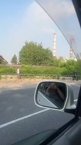 This video is captured from inside a car, looking out the side window and through the windshield. In the foreground, the side mirror of the car is visible, reflecting a person holding a phone, likely taking a selfie. Outside the car, there's a road with a white line marking the lane. Beyond the road, there's a low fence and dense green vegetation. In the background, two tall industrial smokestacks are emitting plumes of white smoke into a clear blue sky. A person in a dark shirt is seen walking along the sidewalk next to the fence. As the video progresses, the view shifts to looking through the windshield. Several vehicles are on the road ahead, including a large white and red bus, a black SUV, and a green sedan. There are also trees and utility poles visible along the side of the road, suggesting a semi-urban or industrial area. The overall scene depicts a journey through an area with industrial activity and road traffic.
