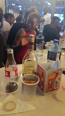 The video shows a crowded outdoor event, possibly a bar or club, with loud music playing. The scene includes multiple tables scattered throughout the space, each adorned with bottles, drinks, and plastic cups. Several people are present, including a woman in a black dress with a silver necklace, and a child in a red dress. The event appears to be lively and festive, with visible vapor or fog enhancing the atmosphere. The background includes string lights and green neon accents, suggesting a modern or party setting.