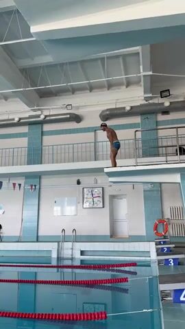 A man in blue swim trunks stands on a diving board above a swimming pool. He jumps off the board and performs a somersault in the air before entering the water. The pool has lane markers and flags are hung along the wall.