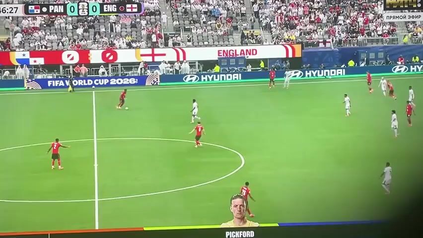 The video shows a soccer match between Panama and England. The score is 0-0. The game is being played on a green grass field with white lines marking the boundaries and a center circle. There are two goal nets visible. Spectators are in the stands surrounding the field. Several players in red and white uniforms are on the field. A player in a red uniform is kicking a soccer ball towards the center of the field. Another player in a white uniform is running towards the ball. A scoreboard is visible at the top of the screen, showing the score and the teams' names. There are advertisements around the stadium, including Hyundai and Qatar Airways. There are also images of three soccer players at the bottom of the screen.