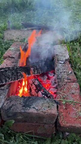 A fire burns in a makeshift brick fire pit. Sparks fly from the fire as a person uses a stick to poke at the burning logs. The fire pit is surrounded by green grass and some foliage. The scene appears to be outdoors.