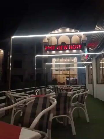 The video shows a nighttime exterior shot of a building with a sign that reads "PINE VIEW HOTEL" in illuminated red letters. The building has a grand entrance with large glass windows and arches. In front of the hotel, there is an outdoor seating area with tables and chairs, some of which are covered with checkered cushions. White string lights are draped across the scene, illuminating the tables and chairs and adding to the ambiance. The overall environment appears to be that of a hotel or restaurant at night.