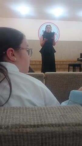 A girl in a black dress and heels stands on a small stage, holding a tablet and reciting a speech. Another girl with glasses, wearing a white shirt, sits in the foreground, facing away from the camera. The background shows a wall with a circular logo and a piano. The scene appears to be an indoor event, possibly a school or concert hall.