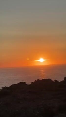 A sunset over the ocean is viewed from a rocky cliff. The sun is low on the horizon, casting a bright orange glow across the sky and the water. A person is visible on the right side of the frame, holding up a phone to take a picture of the sunset. They are wearing a white shirt and are silhouetted against the bright sky. The ocean is calm, with small waves breaking on the shore. The sky is a gradient of orange and yellow, with a few wisps of clouds visible.