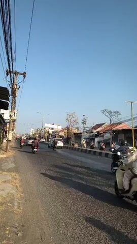 A vertical, eye-level, wide shot shows a busy road on a sunny day. On the left, a dirt path lines the road, with a utility pole covered in wires to the far left. On the right, the road is paved with a rough, dark asphalt. Buildings with red tile roofs line the right side of the road. A line of motorbikes is traveling down the road. The lead motorbike has a person in a red shirt and black pants, the second has a person in a white shirt and blue jeans, and the third has a person in a red helmet and a blue and white shirt. Further down the road, a white van is visible, followed by more motorbikes and a large white truck. The sky is a clear, pale blue.