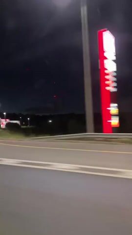 The video shows a view from inside a vehicle at night, driving on a road. The camera is angled to show the road ahead and to the side, as well as the sky. On the left side of the road, there is a brightly lit gas station with a red and white sign. Further down the road, there are streetlights and road signs. A truck is visible on the road, and another truck is seen in the distance. The sky is dark with some lights and faint red specks that could be distant lights or fireworks. The video is shaky, indicating movement.