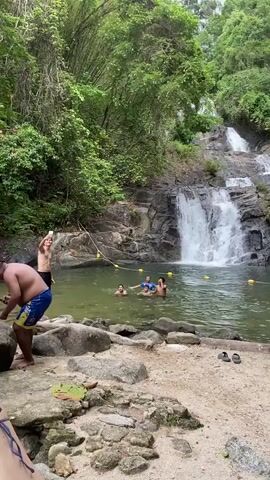 The video depicts a scenic waterfall scene in a lush, green environment. Several people are enjoying the water in a natural pool at the base of the waterfall. One individual is standing on the rocky bank taking a photo, while others are swimming and interacting in the water. The waterfall itself cascades down a rocky slope, and the surrounding area is dense with trees and vegetation. The sky appears overcast. There is some evidence of human intervention, with a rope and yellow buoys floating in the water to delineate a swimming area.