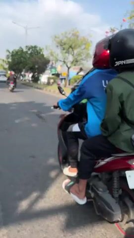 A motorcyclist wearing a black helmet and a green jacket is riding a scooter on a paved road. The scooter has a license plate that reads H 6029 UCC. A black minivan is driving next to the scooter. The minivan has a spoiler and tinted windows. There are trees and buildings in the background. The sky is blue and there are some clouds. The road is worn and has some cracks.