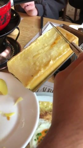 A person is scraping melted cheese from a raclette grill onto a white plate with slices of cooked potato. The cheese is golden and bubbly, and a small knife is being used to scrape it off. In the background, a red pot sits on a stand, likely containing more cheese or a fondue. Another plate with an egg and chives can be seen. The table is wooden and has a newspaper or placemat underneath. The person's hand holding the knife and another person's hand resting on the table are visible, suggesting a group meal.