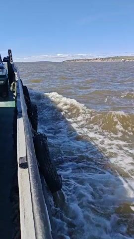 The video shows a view from the side of a moving boat. The water is choppy and creating a wake behind the boat. There are four tires hanging over the side of the boat, which are likely used as fenders. In the distance, there is a shoreline with some hills or cliffs. The sky is blue and clear. The video is shaky, indicating that the boat is in motion.