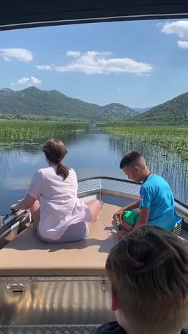 In this video, three people are on a boat, two of whom are visible from the back, sitting on the bow. The person on the left, a woman, is wearing a light pink t-shirt and grey shorts. The person on the right, a boy, is wearing a blue t-shirt and green shorts. A third person, whose back is towards the camera, is in the foreground, obscuring the lower part of the boat.

They are on a calm body of water, likely a lake, surrounded by lush green hills and mountains. Tall reeds and lily pads are visible in the water, creating a natural and serene environment. The sky is a clear blue with a few scattered white clouds, suggesting a pleasant, sunny day. The light indicates it is daytime, likely midday or early afternoon.

The boat appears to be moving slowly through the water, as suggested by the slight ripple. The boy on the right is interacting with something on his lap, possibly reeds or a small plant. The overall mood seems peaceful and relaxed, perhaps a scenic boat tour.

The location is identified as Prevlaka, Montenegro. No visible text is present in the video. The boat itself has a metallic hull with some visible latches and a tan-colored padded seating area. The scene is dominated by the natural landscape and the tranquil atmosphere of the boat ride.