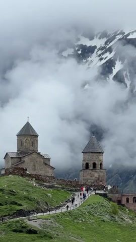 The video shows a view of a stone church and bell tower on a green grassy hill. Several people are walking along a path leading to the church. The sky is overcast with clouds, and a snow-capped mountain is visible in the background, partially obscured by fog. The church has a conical roof topped with a cross, and the bell tower also has a conical roof. The overall scene is one of a remote, mountainous location with a historical religious structure.